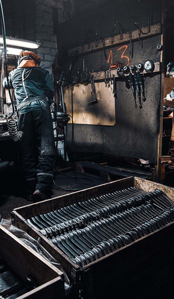 At the dark workshop experienced worker in protective uniform is working with metal.